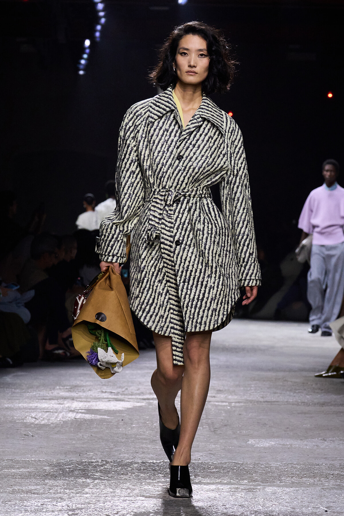 Female model walking a runway in a black-and-white striped belted coat, carrying a brown paper bag with flowers inside.] repeated?