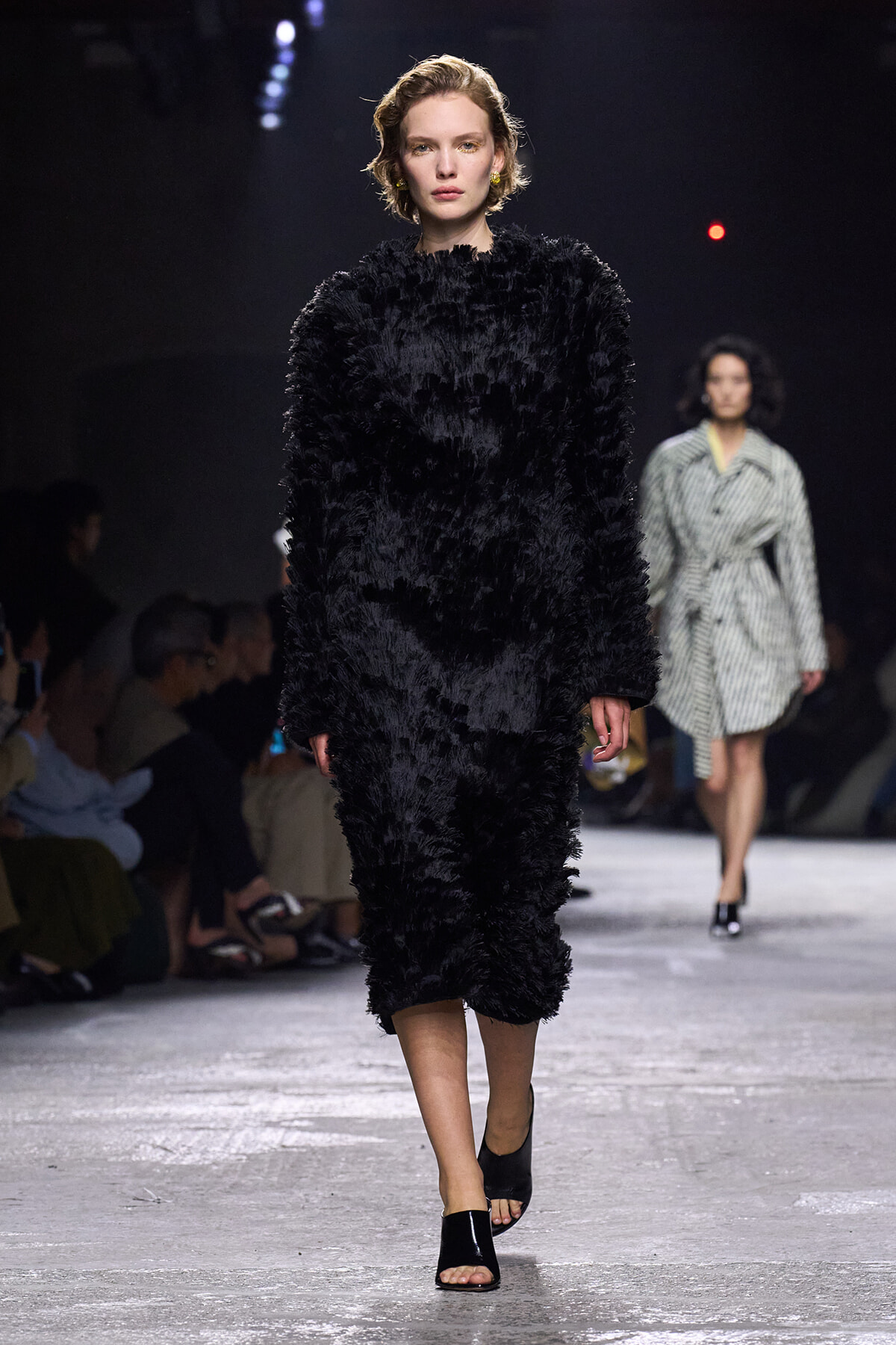 Model walking the runway in a voluminous black fluffy coat, with a stern expression, as another model in a striped coat follows in the background. Audience seated along the catwalk on the left.