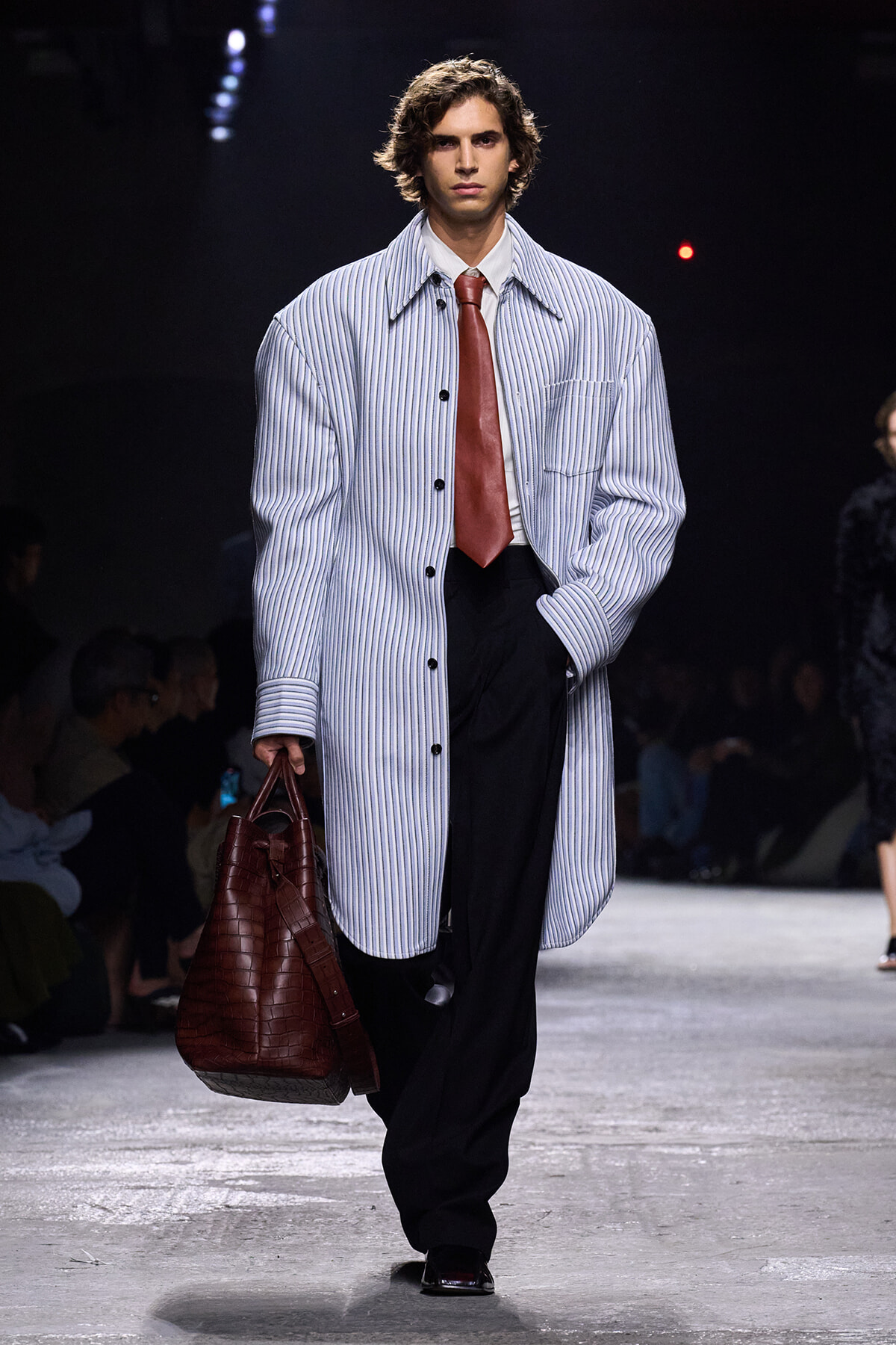 Male model walking a fashion runway wearing a long light-blue striped coat, white shirt, red tie, black trousers, and carrying a large burgundy crocodile‑patterned bag.