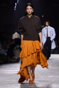 Model on a fashion runway wearing a dark brown oversized V-neck sweater and a bright orange tiered ruffled skirt with orange woven sandals.