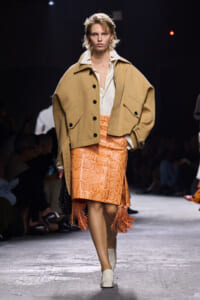 Runway model wearing a camel oversized jacket over a white shirt and a bright orange snake-skin skirt with fringe at the hem.