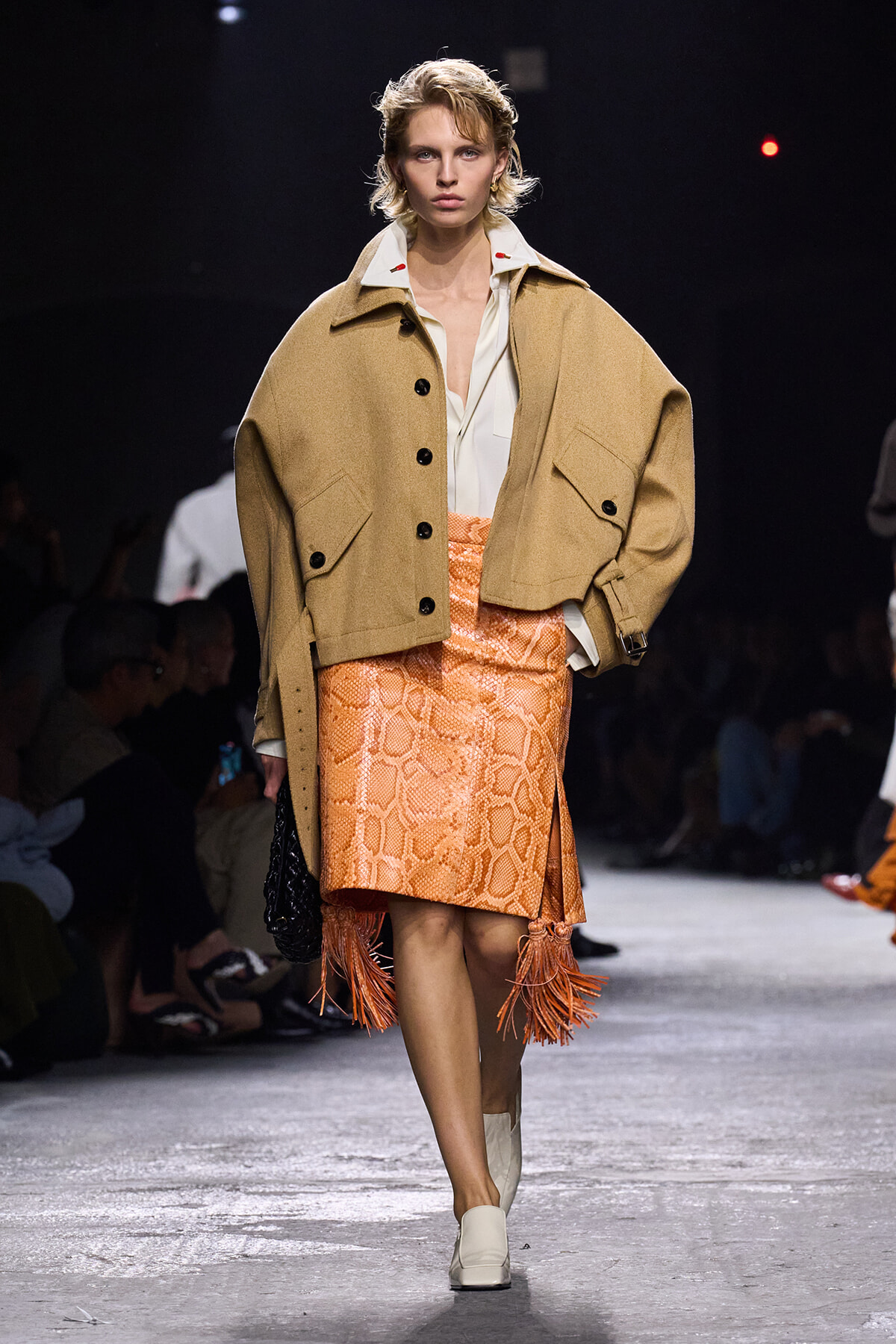 Runway model wearing a camel oversized jacket over a white shirt and a bright orange snake-skin skirt with fringe at the hem.