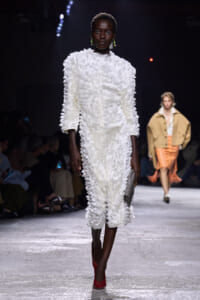 Model walks the runway in a white feathered midi dress, red heels, and a gray clutch on a dark backdrop.