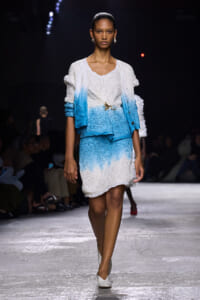 Model walks a runway wearing a white-to-blue gradient knit dress with matching light cardigan and a gold brooch at the waist, white heels.