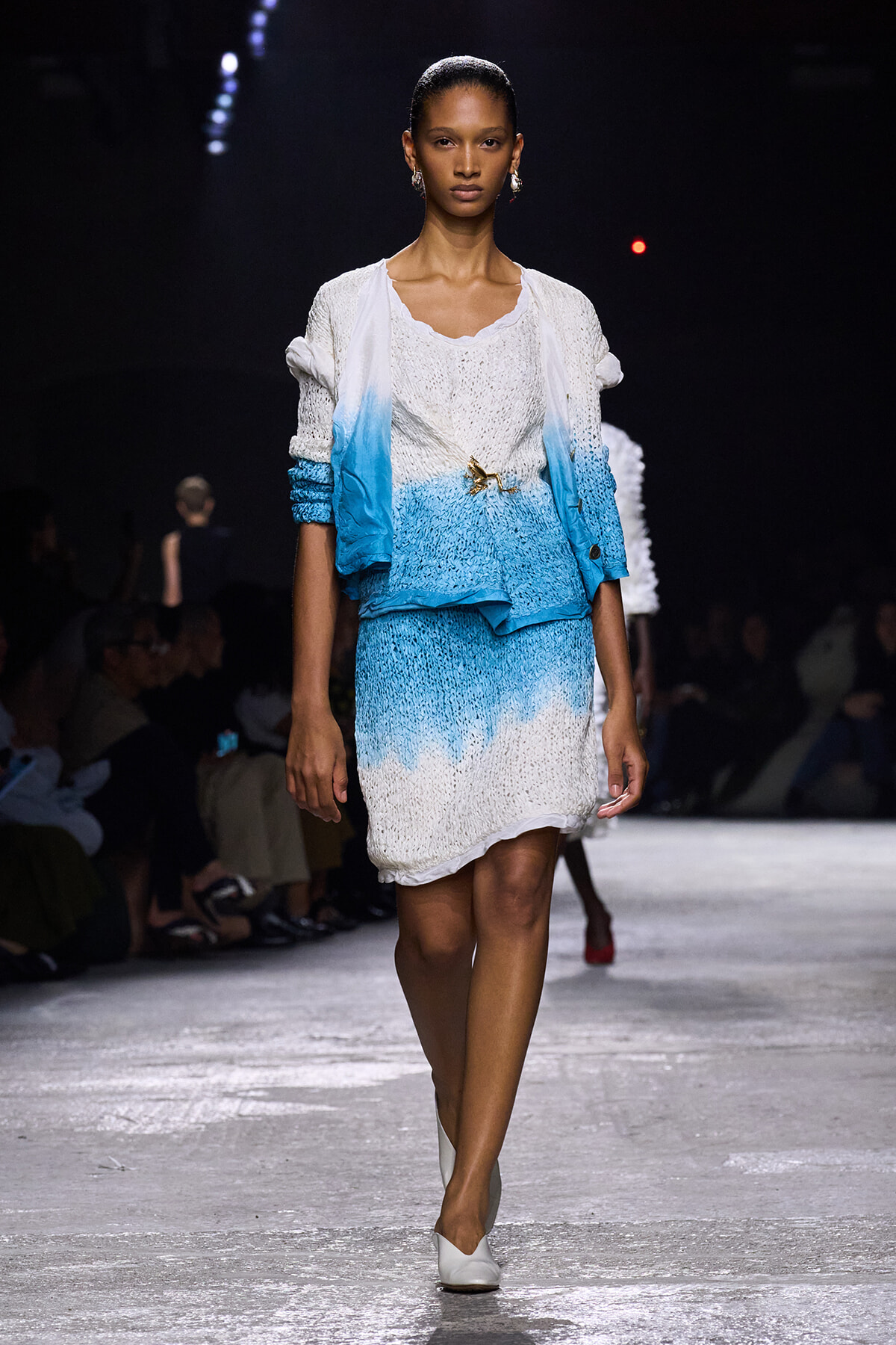 Model walks a runway wearing a white-to-blue gradient knit dress with matching light cardigan and a gold brooch at the waist, white heels.