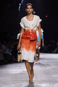 Model walks the runway in a cream-to-orange gradient knit dress with rolled sleeves, carrying a straw handbag.
