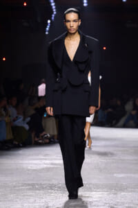 Model walks the runway in a black, oversized blazer with sculptural, circular lapels and matching trousers.