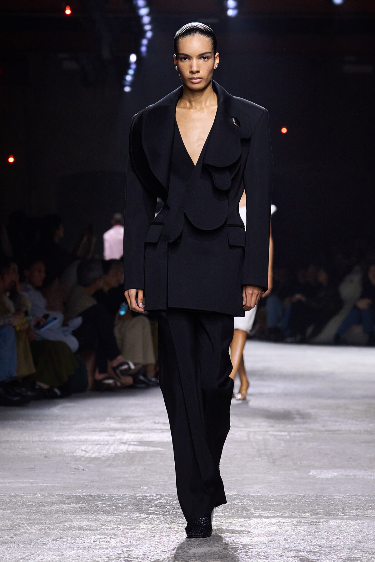 Model walks the runway in a black, oversized blazer with sculptural, circular lapels and matching trousers.