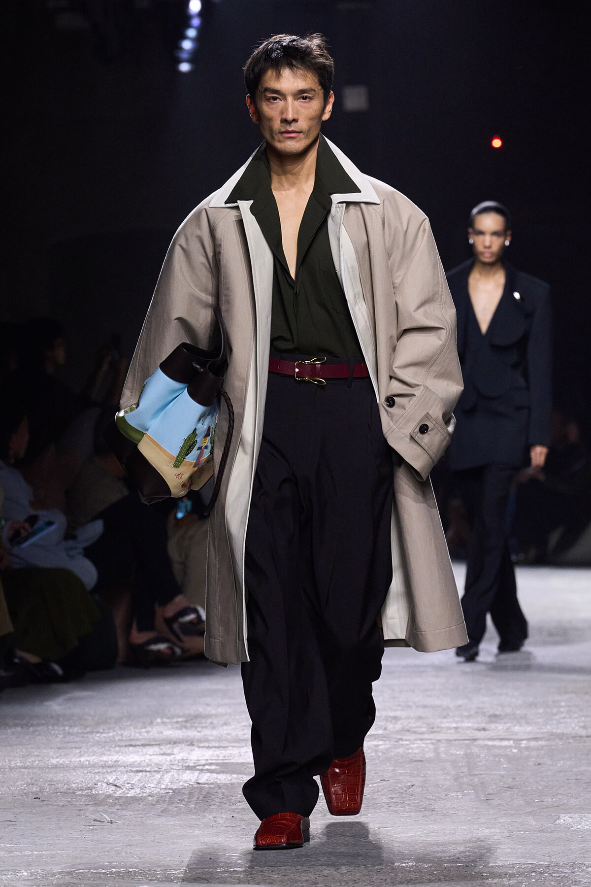 Male model walks a fashion runway in a beige trench over a dark green shirt and black pleated pants, with a red belt, red boots, and a colorful tote.