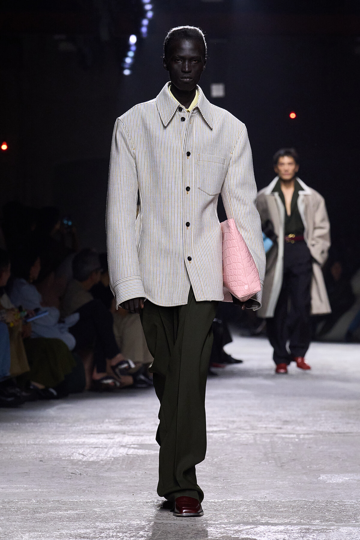 Male runway model wearing a light striped button-up jacket with oversized collar, olive wide-leg pants, and a pink quilted handbag on the arm.