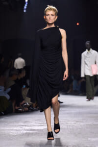 Model walks the runway in a black one-shoulder draped midi dress with a cape-like sleeve and gold earrings.