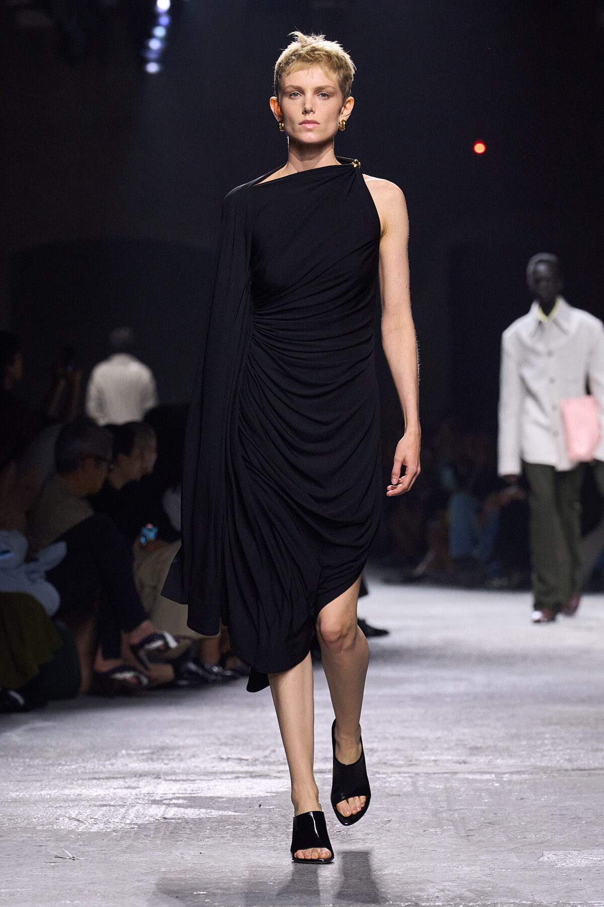 Model walks the runway in a black one-shoulder draped midi dress with a cape-like sleeve and gold earrings.