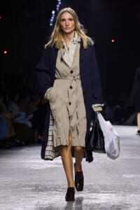 Female model walks a fashion runway in a crinkled beige trench dress with a navy overcoat, carrying a black handbag and a white shopping bag.