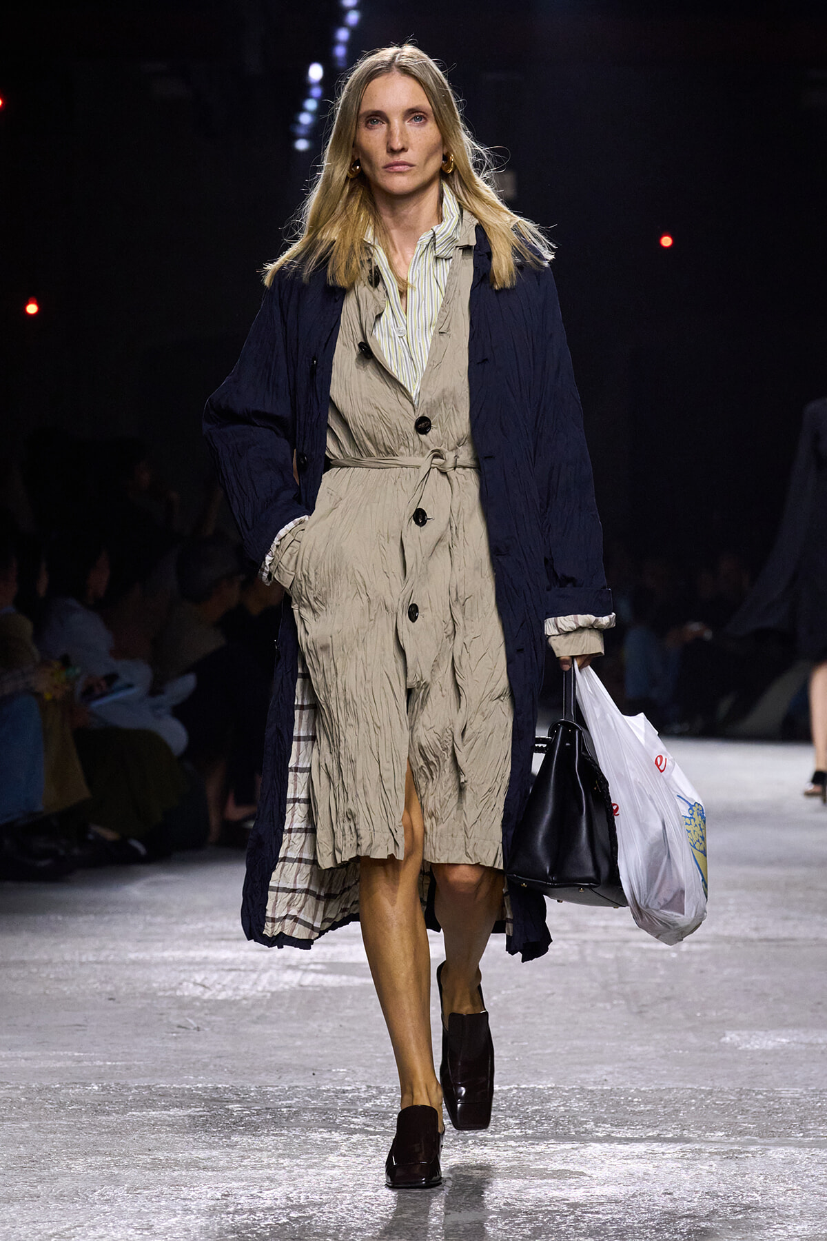 Female model walks a fashion runway in a crinkled beige trench dress with a navy overcoat, carrying a black handbag and a white shopping bag.