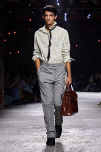 Male model on a runway wearing a striped button-up shirt, crinkled light gray trousers, and a dark scarf, carrying a brown textured bag.