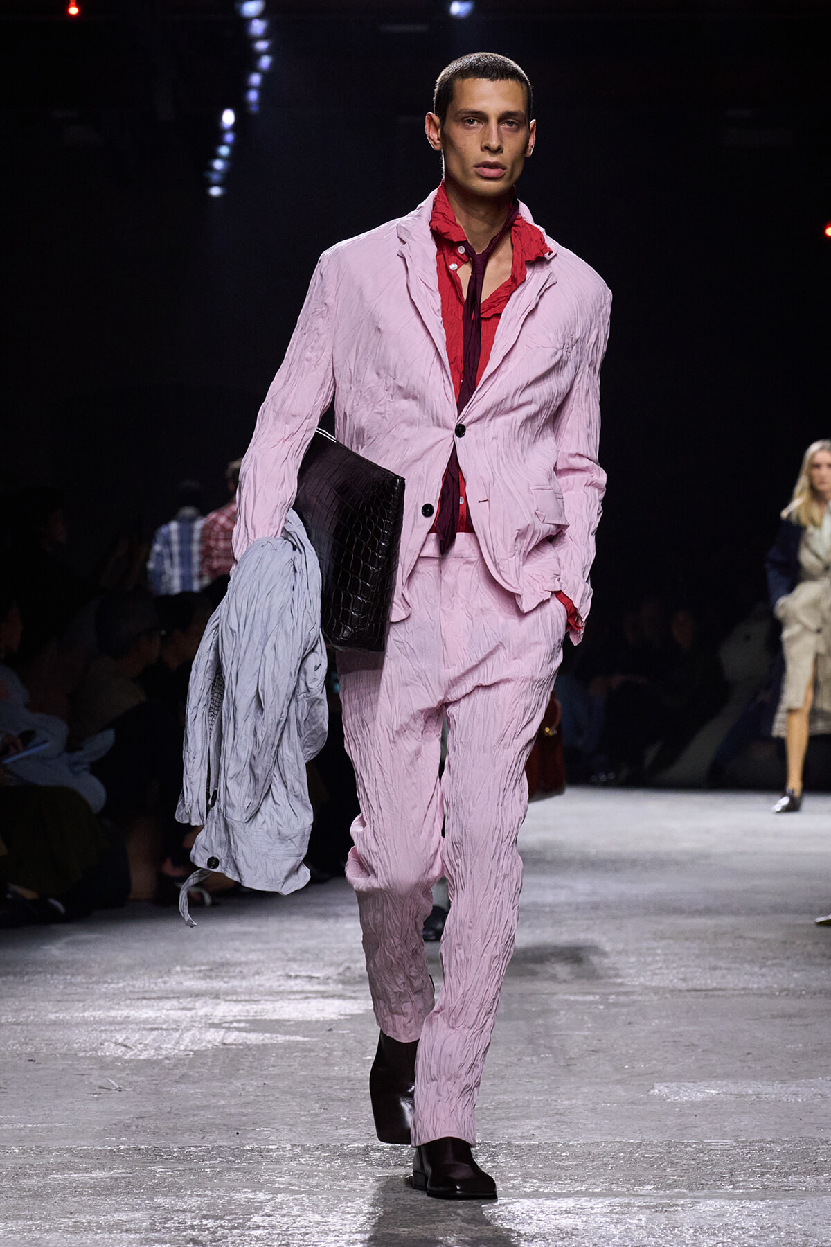 Male model walks a fashion show runway in a crinkled pink suit with a red shirt, carrying a gray jacket and a black portfolio clutch.