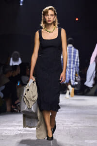 Model walking a runway in a black textured midi dress with a chunky green bead necklace, carrying a beige coat and gray handbag.