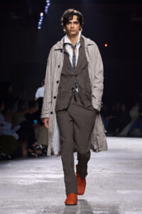 Male model on a runway wearing a layered crinkled gray-brown suit with a light beige trench and bright orange textured boots.