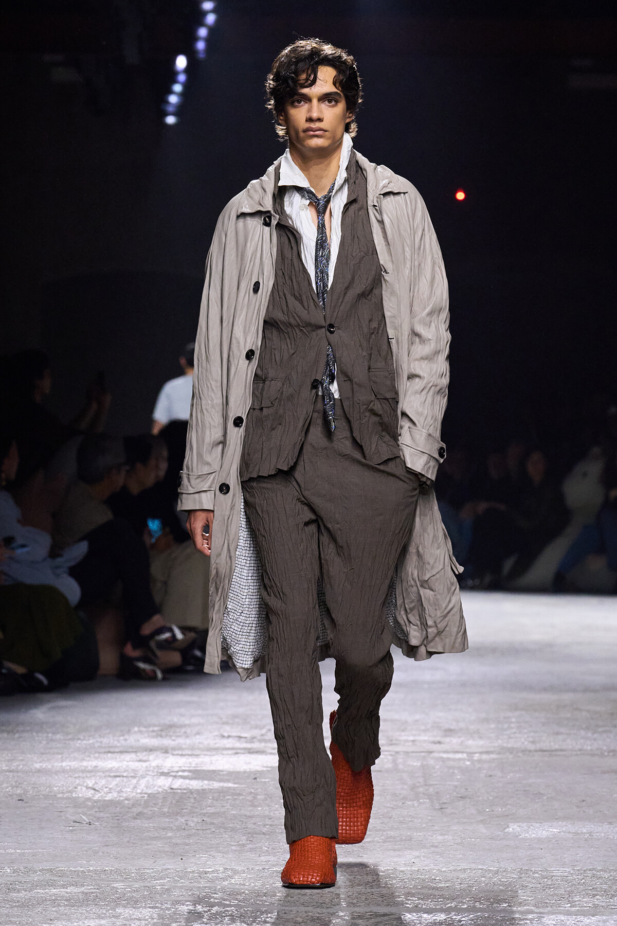 Male model on a runway wearing a layered crinkled gray-brown suit with a light beige trench and bright orange textured boots.