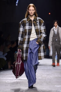 Model walking a runway in an oversized plaid coat over a cream knit, with wide-leg blue pants and a burgundy stitched tote bag in hand