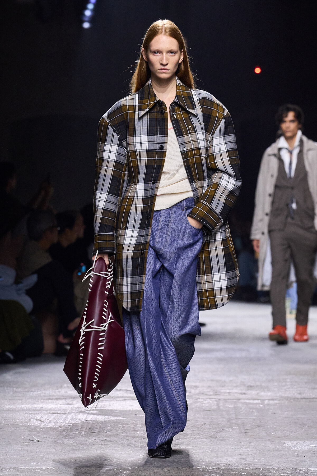 Model walking a runway in an oversized plaid coat over a cream knit, with wide-leg blue pants and a burgundy stitched tote bag in hand