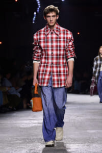 Male model on a runway wearing a red plaid oversized shirt, blue wide-leg pants, white sneakers, and an orange woven bag.