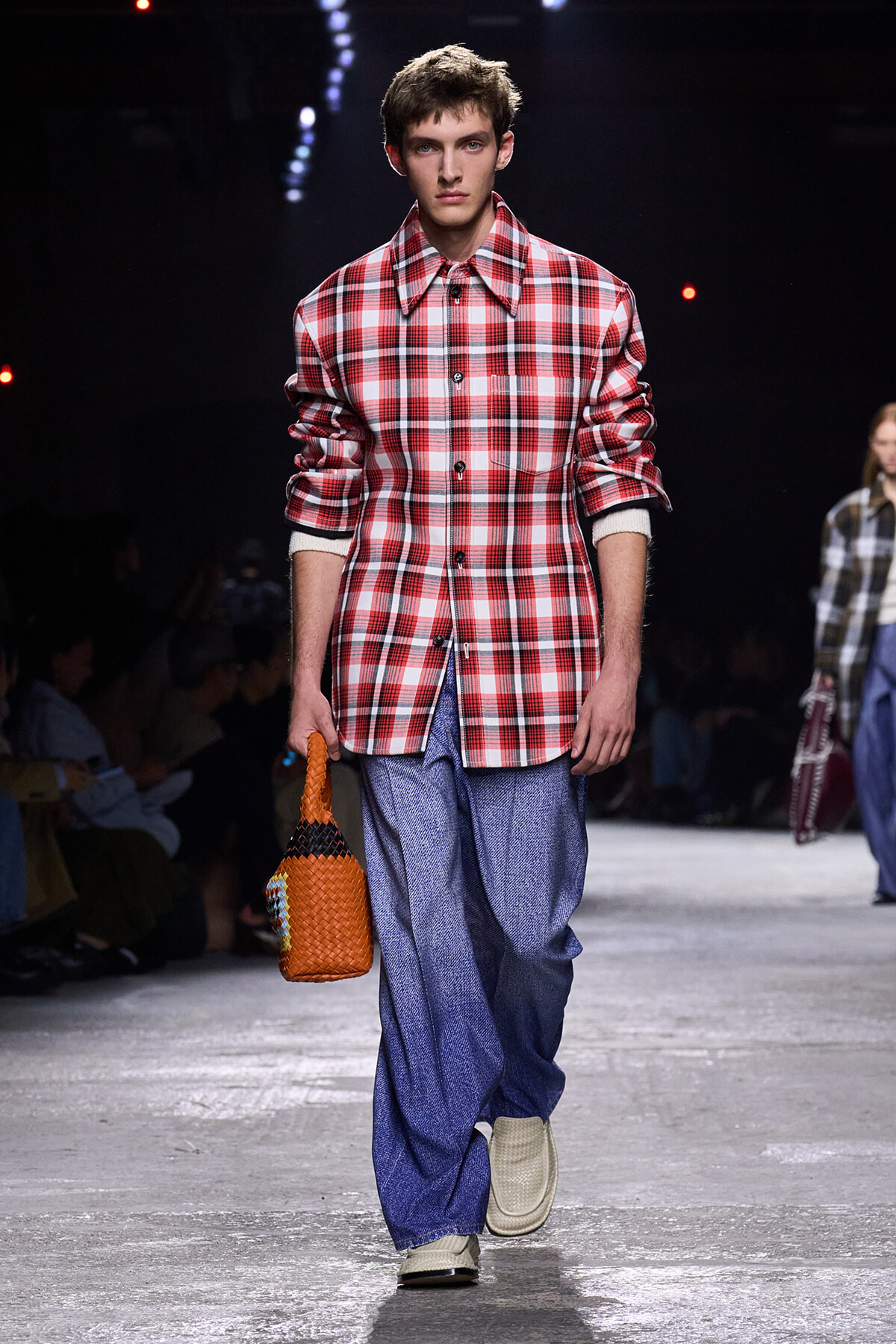 Male model on a runway wearing a red plaid oversized shirt, blue wide-leg pants, white sneakers, and an orange woven bag.