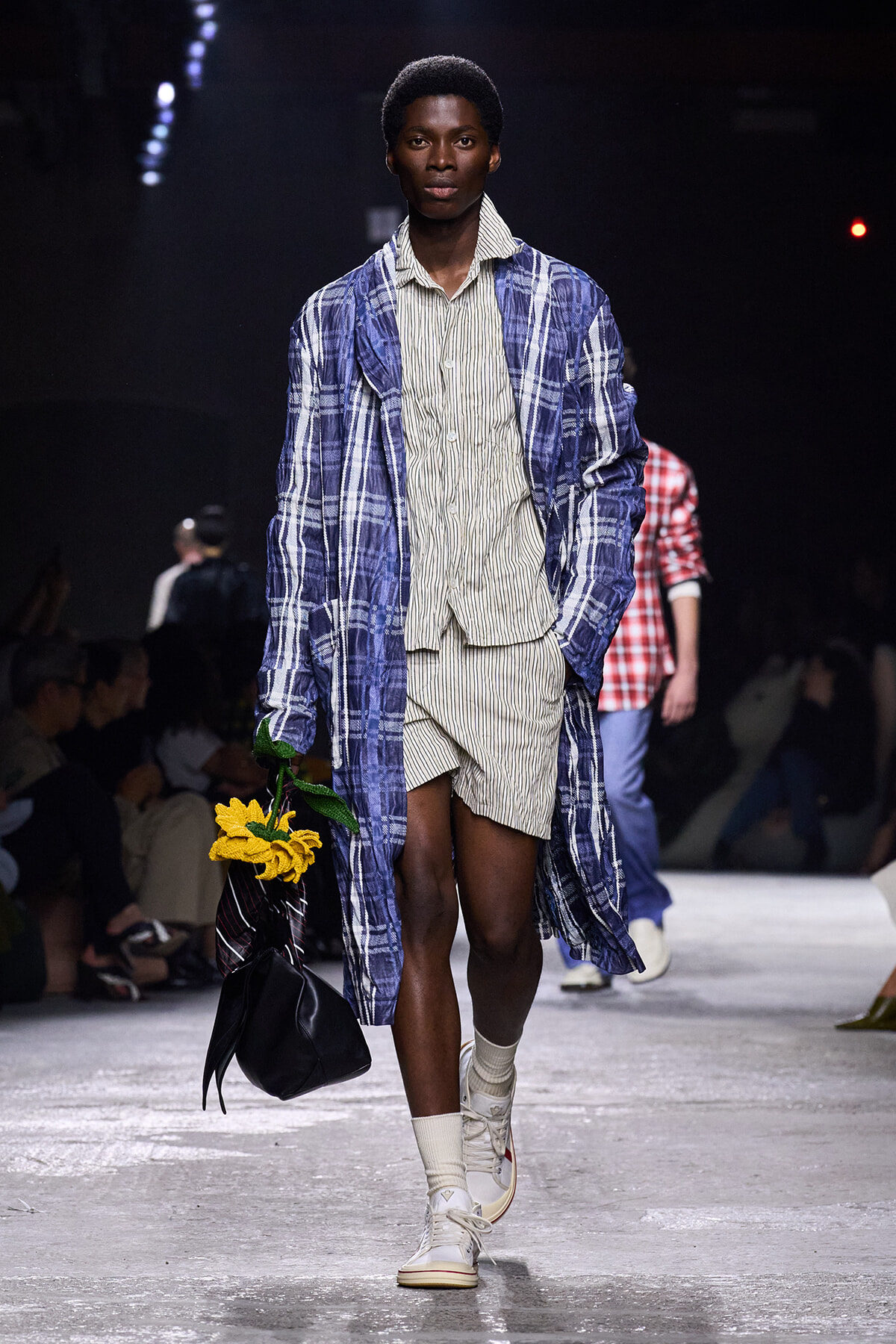 Male model walking a fashion runway in a blue plaid long coat over beige striped shirt and shorts, carrying a black handbag with sunflowers.