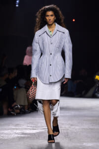 Model walking a fashion show in a blue-and-white striped oversized button-front coat, mint blouse, and white fringe-hem skirt, carrying a woven red-and-cream bag.