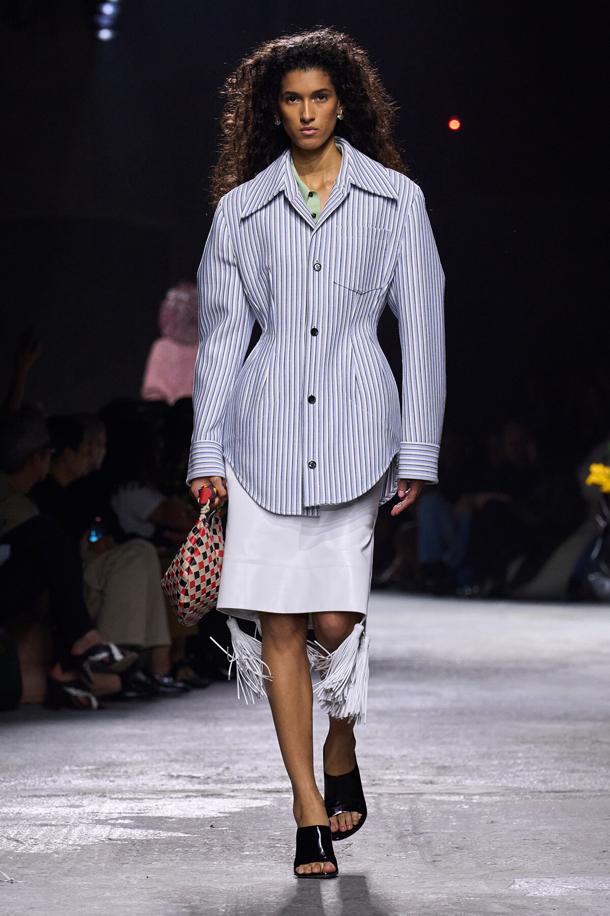 Model walking a fashion show in a blue-and-white striped oversized button-front coat, mint blouse, and white fringe-hem skirt, carrying a woven red-and-cream bag.