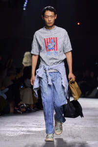 Male model walks a fashion show wearing a grey graphic T-shirt with red stripes and rabbit print, a striped shirt tied around the waist, light denim pants, sneakers, and carries two bags.