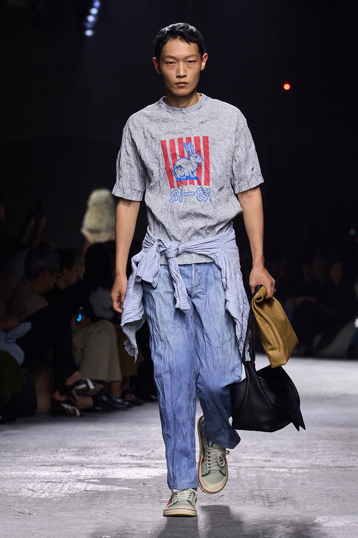 Male model walks a fashion show wearing a grey graphic T-shirt with red stripes and rabbit print, a striped shirt tied around the waist, light denim pants, sneakers, and carries two bags.