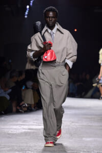 Male model walking a runway in an oversized beige trench, gray tie, and wide beige trousers, carrying a red drawstring bag and a black quilted backpack while wearing red sneakers.