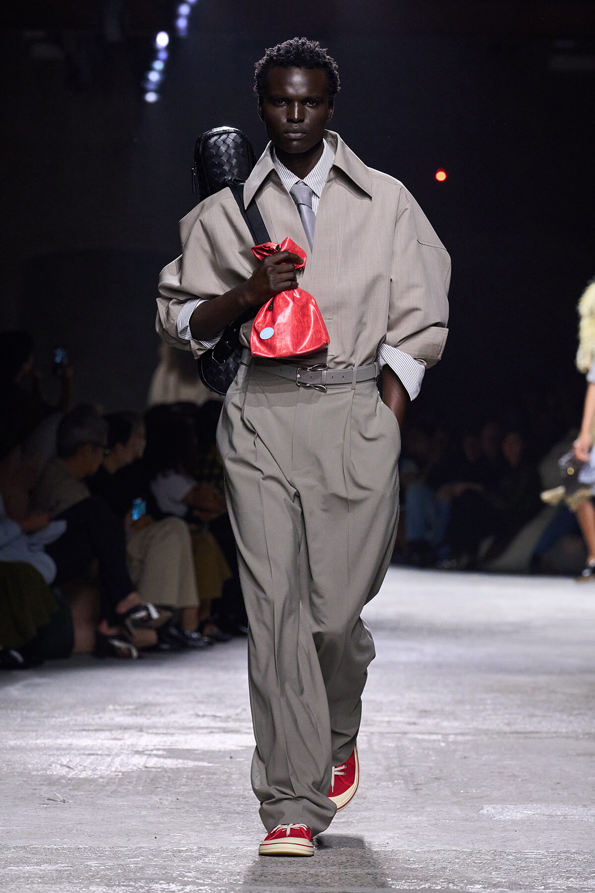 Male model walking a runway in an oversized beige trench, gray tie, and wide beige trousers, carrying a red drawstring bag and a black quilted backpack while wearing red sneakers.