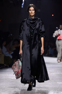 Runway model in a black satin draped coat with voluminous sleeves, carrying a multicolored fringed handbag.