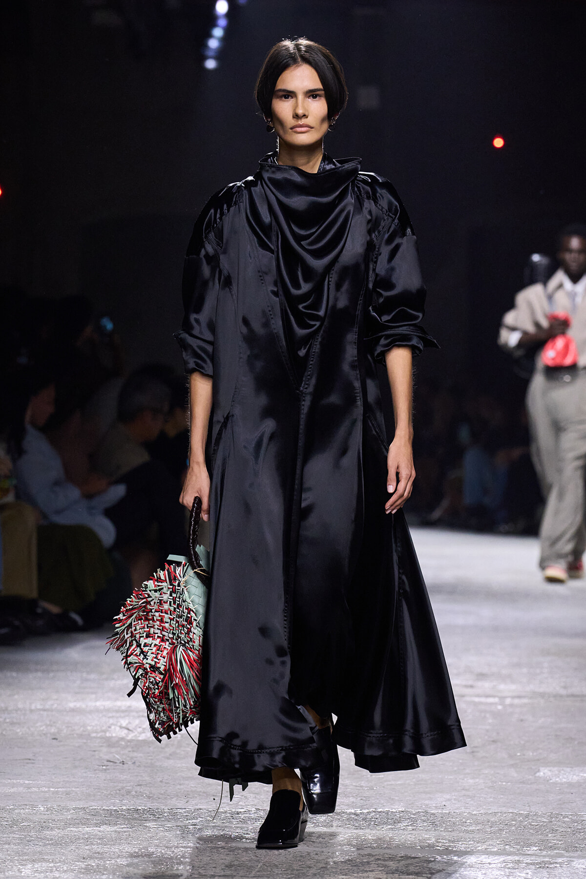 Runway model in a black satin draped coat with voluminous sleeves, carrying a multicolored fringed handbag.