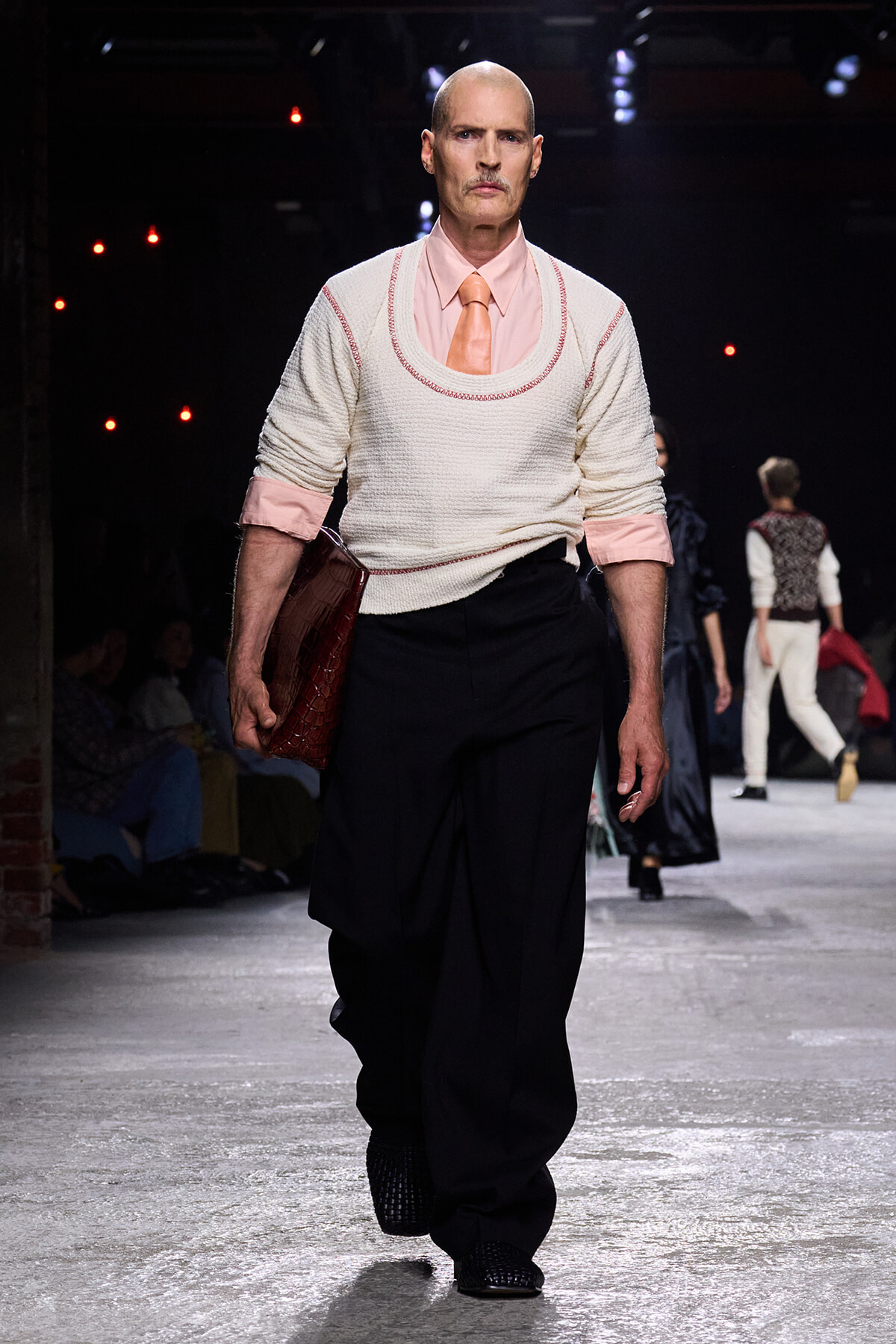 Male model walking a fashion runway in a pale pink shirt, peach tie, white sweater vest with pink trim, and black wide-leg pants, carrying a red clutch.