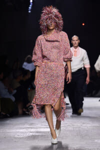Model walking a fashion show in a speckled pink knit dress with fringe tassels and a spiky feathered hood for the look's dramatic effect.