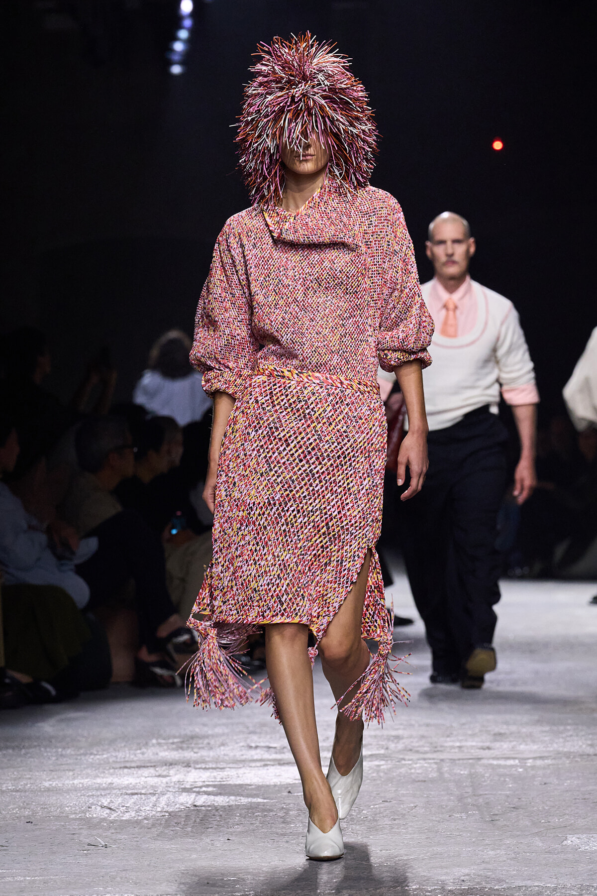 Model walking a fashion show in a speckled pink knit dress with fringe tassels and a spiky feathered hood for the look's dramatic effect.