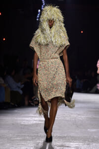 Model on a runway wearing a beige textured dress with red speckles and a dramatic fringed hood made of straw-like fibers, holding a dark clutch.