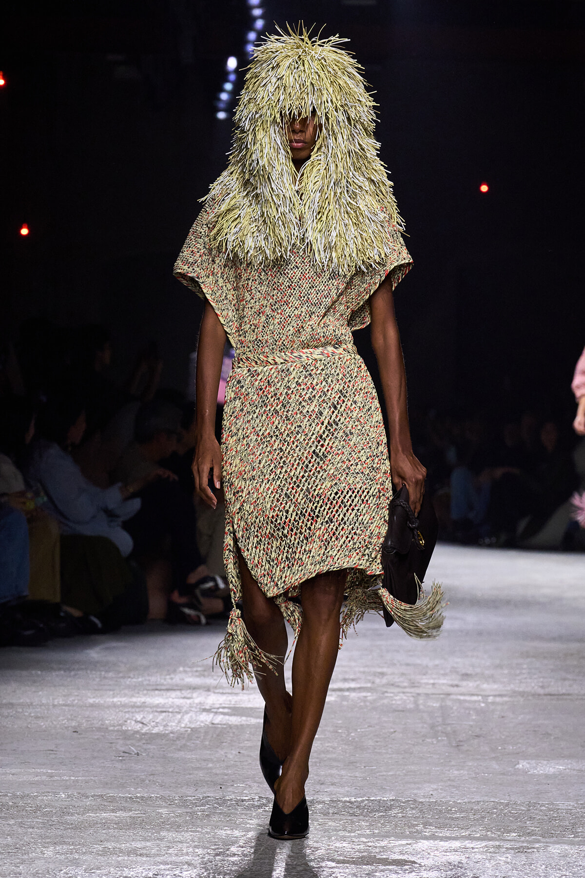 Model on a runway wearing a beige textured dress with red speckles and a dramatic fringed hood made of straw-like fibers, holding a dark clutch.