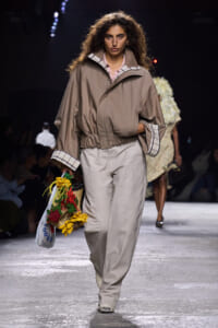 Model walking a runway in a beige oversized jacket with plaid cuffs and wide tan pants, carrying a bouquet of sunflowers and a plastic tote.