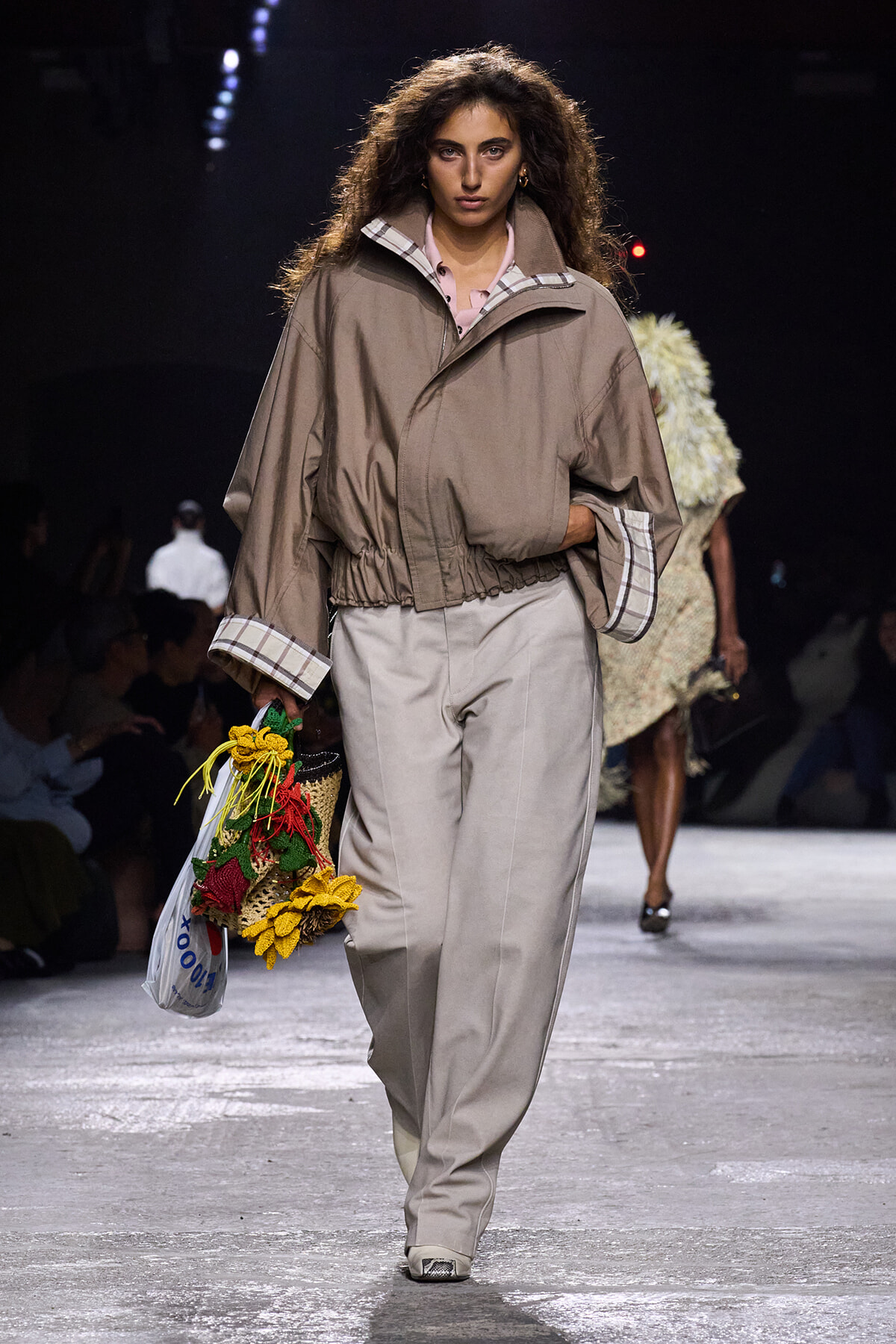 Model walking a runway in a beige oversized jacket with plaid cuffs and wide tan pants, carrying a bouquet of sunflowers and a plastic tote.