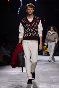 Male model walking a fashion runway in a white knit outfit with a maroon-patterned sleeveless sweater, carrying a red coat and a black woven backpack.
