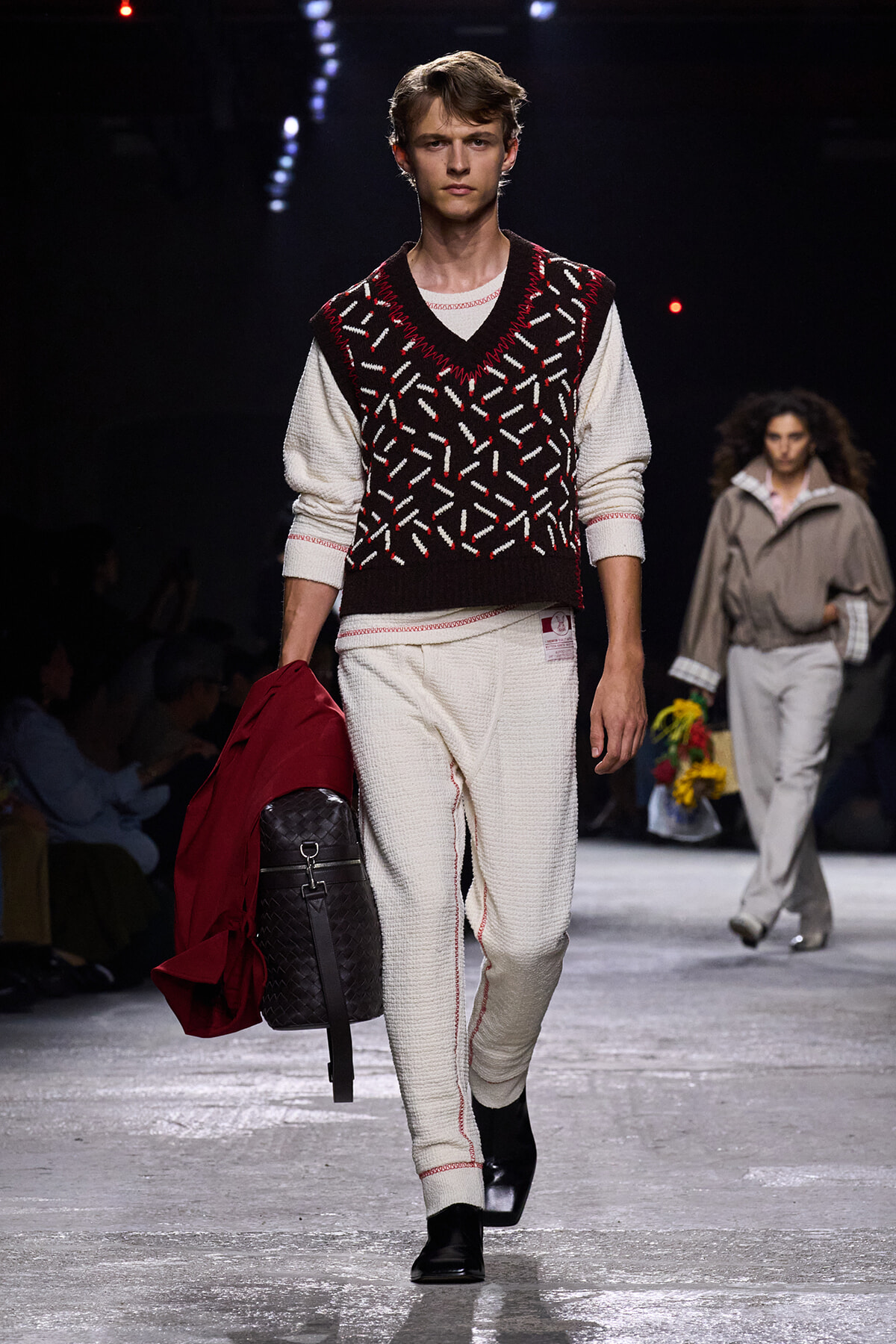 Male model walking a fashion runway in a white knit outfit with a maroon-patterned sleeveless sweater, carrying a red coat and a black woven backpack.