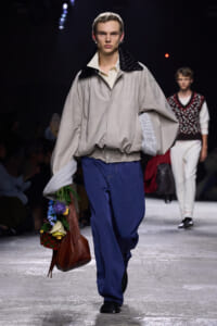 Male model on a runway wearing an oversized beige jacket with a black quilted collar and blue wide-leg pants, carrying a brown handbag overflowing with colorful flowers.