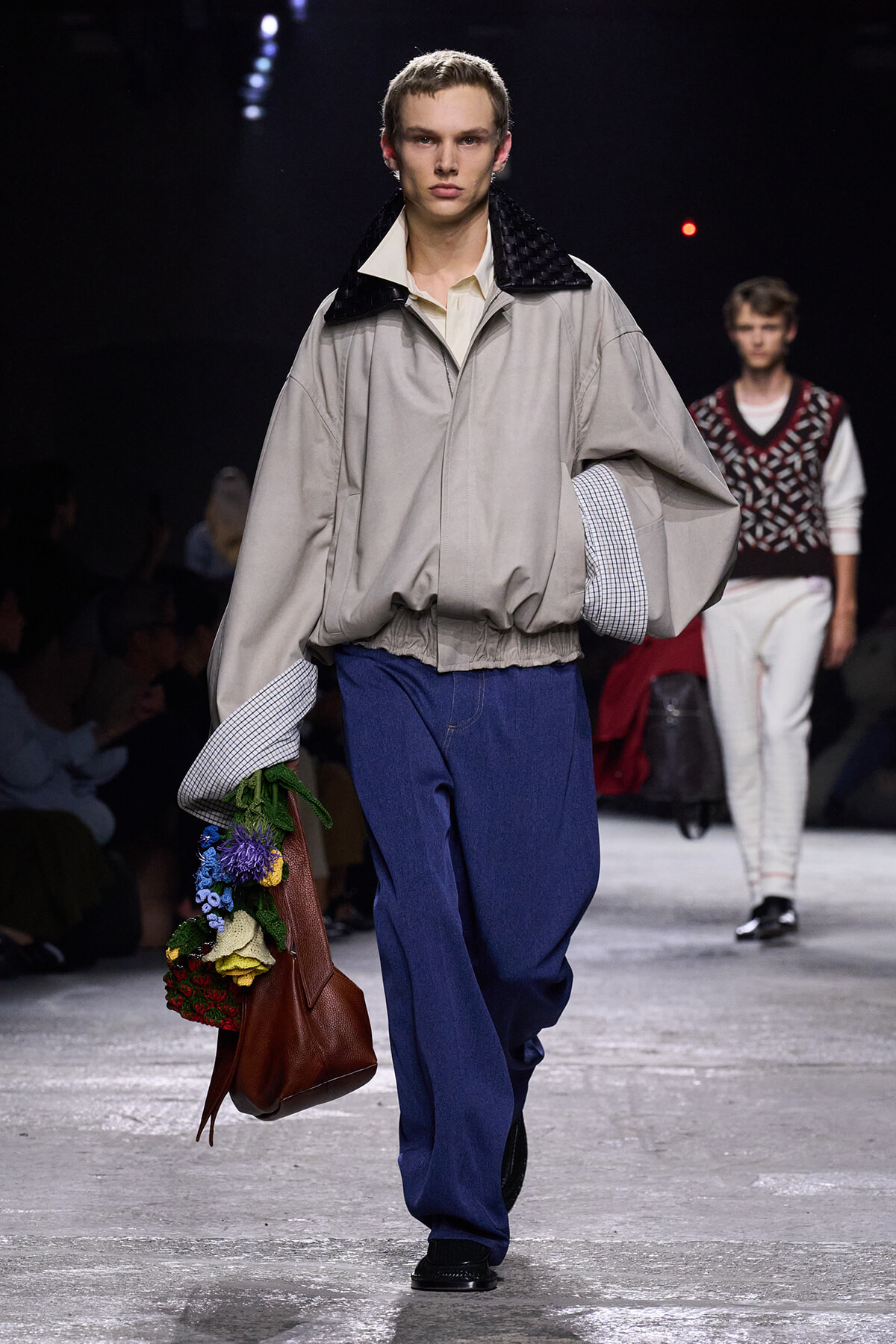 Male model on a runway wearing an oversized beige jacket with a black quilted collar and blue wide-leg pants, carrying a brown handbag overflowing with colorful flowers.