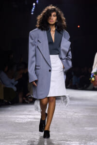 Model walks a runway in an oversized light gray blazer over a dark teal blouse and a white fringe-hem skirt, black heels, and voluminous curls.