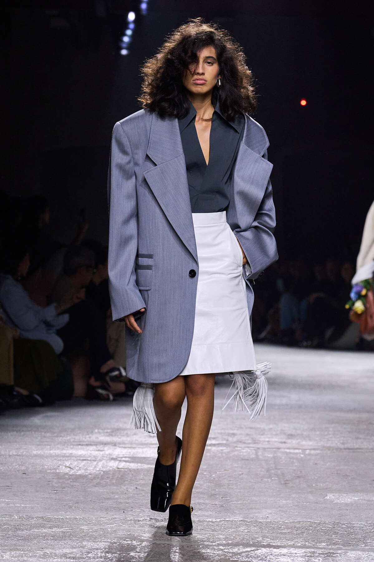 Model walks a runway in an oversized light gray blazer over a dark teal blouse and a white fringe-hem skirt, black heels, and voluminous curls.