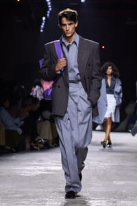Male model walks the runway in an oversized gray suit with a light blue shirt, striped tie, and a purple shoulder bag.